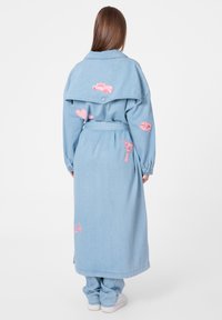 Manteau long en denim bleu clair avec des détails brodés roses, comprenant des cœurs et des voitures, et une ceinture assortie. Manches longues et coupe décontractée.