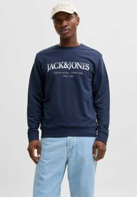 Marineblå sweatshirt med hvid "JACK&JONES" tekst, rund halsudskæring, ribbede manchetter og kant, parret med lyseblå denimbukser.