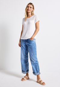 Maglietta bianca con un design a rete aperta e testurizzato nella parte superiore. Abbinata a jeans larghi blu chiaro e sandali a scivolo beige.