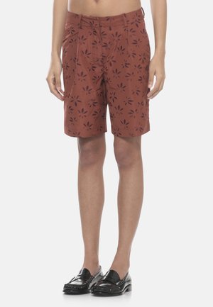 Gambe snelle che indossano pantaloncini color ruggine con stampe floreali scure e mocassini in pelle nera slip-on, in piedi su uno sfondo chiaro.