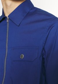 Camisa azul de tela suave, con cuello puntiagudo, cremallera plateada y un bolsillo en el pecho con un acento de botón redondo.
