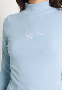 Maglione a collo alto a coste di colore azzurro chiaro con maniche lunghe, dotato di un dettaglio ricamato bianco con firma sul petto. Design morbido e aderente.