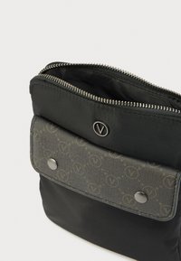 Črna tkaninska torbica z zgornjo preklopno zanko, ki ima vzorčni logotip in dva kovinska potisna gumba, na vrhu pa srebrn zadrga.