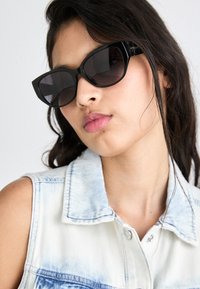 Schwarze Cateye-Sonnenbrille mit dunklen Gläsern, glänzender Oberfläche und dezenten Details an den Bügeln. Getragen mit einer leichten Jeansweste.