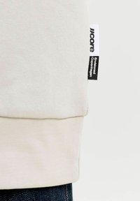 Tissu beige avec deux étiquettes cousues sur la couture, une blanche avec un texte sombre « core », et une noire avec un texte blanc « Envisioned Copenhagen ».