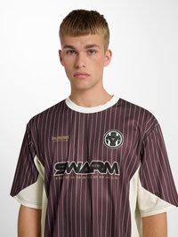Junger Mann mit kurzen Haaren, der ein braunes Sporttrikot mit Nadelstreifen und der Aufschrift „SWARM INTERNATIONALS“ sowie Logos auf weißem Hintergrund trägt.
