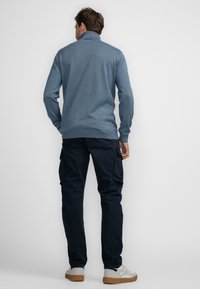 Blauwe gebreide trui met een hoge kraag, gecombineerd met marineblauwe cargobroek en lichte sneakers. Heeft een ontspannen pasvorm en geribbelde manchetten.