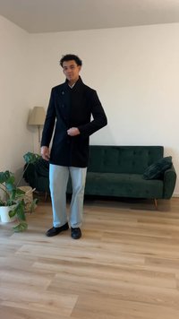 Must häbemust villane mantel kõrge kraega ja kahe ridadega nööpidega, kantud helesiniste pükste ja mustade kingade peal, modernses sisekujunduses.