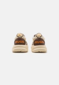 Sneakers w beżowo-brązowym schemacie kolorystycznym, z siateczki i materiału syntetycznego, z wyraźnym logo "HUGO" na pięcie oraz wyściełanym kołnierzem.