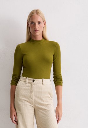 Blonde vrouw met steil haar, gekleed in een olijfgroene longsleeve en hoog-gepaste crèmekleurige ribfluwelen broek, staand tegen een witte muur.