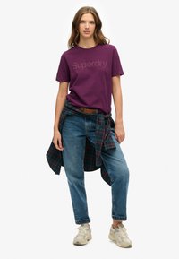 Fioletowa bawełniana koszulka z logo "Superdry", zestawiona z wysokim stanem niebieskimi dżinsami oraz koszulą w kratę zawiązaną w talii. Białe trampki.