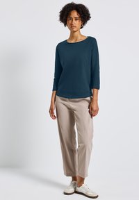 Bluzka z długim rękawem w odcieniu teal z dekoltem łódkowym, zestawiona z jasnobeżowymi spodniami o szerokich nogawkach. Stylizacja uzupełniają casualowe białe sneakersy.