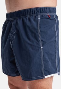 Navy swim shorts made of lightweight fabric feature an elastic waistband, white stitching, and a small red accent on the front.
