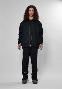 Set di felpa e pantaloni tuta nera, caratterizzato da una parte frontale con zip, due tasche anteriori e tessuto strutturato. Scarpe nere con lacci completano l'outfit.