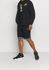Schwarze Windjacke mit "LAKERS" in Gelb, schwarzes T-Shirt mit Grafikdruck, schwarze Shorts mit gemustertem Saum und schwarze Sportschuhe.