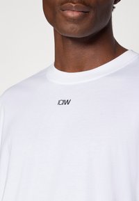 Άσπρο βαμβακερό t-shirt με στρογγυλή λαιμόκοψη και κοντά μανίκια· τυπωμένο λογότυπο "ICIW" σε μαύρο στην αριστερή πάνω πλευρά του στήθους.