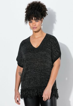 Vrouw met krullend haar die een zwart korte-mouwen gebreide top met franjerand en zwarte leren broek draagt, staand tegen een witte achtergrond.