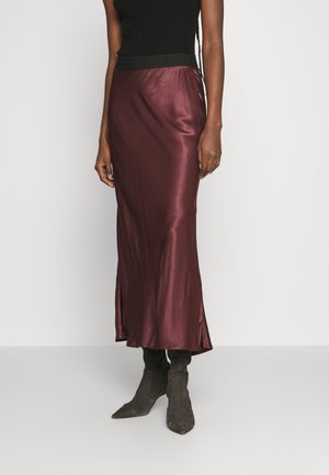 Femme portant un haut noir sans manches, une jupe midi en satin bordeaux brillant et des bottes en suède noires à bout pointu, debout devant un fond uni.