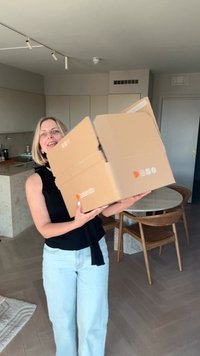 Barna kartondoboz narancssárga logóval, mindkét kézben tartva. Belső tér világos fa padlóval és egy kerek, márvány tetejű asztallal.