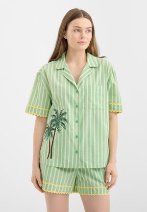 Ung kvinde iført grøn og cremefarvet kortærmet top med striber og shorts med gule, bølgede kanter og palmetræbroderi på toppen.