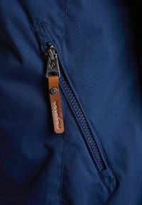 Détail d'une veste bleu marine avec une fermeture éclair en métal ornée d'un tire-zip en cuir marron et d'une languette cousue portant le nom de la marque.