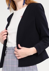 Námořnický žebrovaný blazer s otevřeným předním designem, dlouhými rukávy a strukturovaným střihem. Kombinováno se světlou košilí a kostkovanými kalhotami.