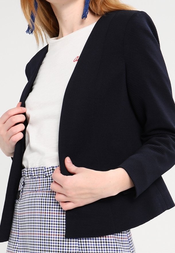 Námořnický žebrovaný blazer s otevřeným předním designem, dlouhými rukávy a strukturovaným střihem. Kombinováno se světlou košilí a kostkovanými kalhotami.