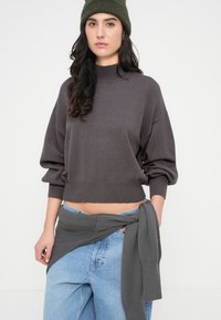 Σκούρο γκρι κοντό πουλόβερ με ψηλό λαιμό, φαρδιά μανίκια και ριμπ τελείωμα, συνδυασμένο με ανοιχτό μπλε τζιν τζιν και ταιριαστό γιλέκο.