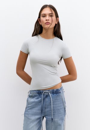 Lichtgrijze, getailleerde top met korte mouwen van rekbaar materiaal, gecombineerd met verouderde lichtblauwe jeans met een trekkoord in de taille.