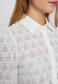 Witte blouse van doorschijnende kant met een bloemenpatroon, knoopsluiting aan de voorkant en klassieke kraag; textuur met subtiele details.