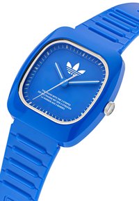 Mėlynas kvadratinis laikrodis su guminiais dirželiu, turintis blizgų paviršių, baltus valandų indikatorius ir „Adidas“ logotipą su tekstūruotu ciferblatu. Sidabrinė karūnėlė.