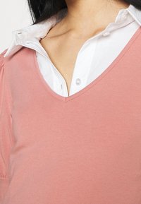Różowy top w serek z miękkiego materiału, nałożony na białą koszulę z kołnierzykiem. Kołnierzyk jest sztywny z detalem guzika przy szyi.