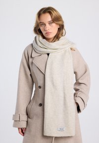 Écharpe en laine beige présentée drapée autour du cou, avec une texture douce et un motif subtil. Étiquette avec logo sur le bord de l'écharpe.