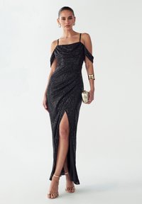 Črna večerna obleka z bleščečimi bleščicami, z naramnicami brez ramen, nabrano zasnovo, razporkom do stegna, dopolnjena z zlatimi dodatki in sandali z zapestnico.