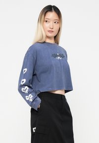 Kratka modra bombažna majica s dolgimi rokavi, ki ima bele cvetno- in srčno grafike na rokavih ter majhen dizajn na prsih.