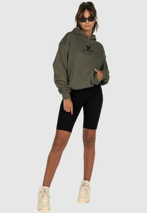 Olivgrüner Hoodie mit einer Vorderasche und schwarzer Schrift, kombiniert mit schwarzen Leggings und beigen High-Top-Sneakern. Die Accessoires umfassen Sonnenbrillen.