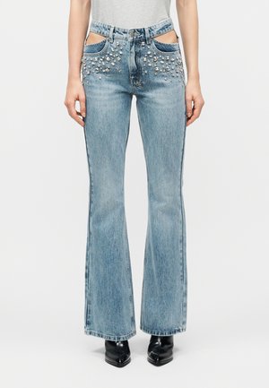 Τζιν με καμπάνες, διακοσμημένα με στρας γύρω από τις μπροστινές τσέπες, φορεμένα με μαύρες αρβυλάκια με μυτερό πάτο και ένα ανοιχτό γκρι πουκάμισο φουσκωμένο μέσα.