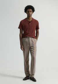 Camiseta henley color burdeos, pantalones claros con patrón marrón y rayas verticales, zapatos marrones. Tejido suave con corte relajado. Líneas limpias.