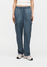 Γυναίκα που φορά μπλε παντελόνι καρό με ελαστική μέση και λευκά αθλητικά παπούτσια, στέκεται με τα χέρια χαλαρά κατά μήκος του σώματος σε απλό φόντο.