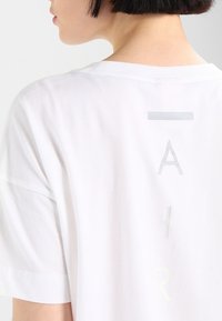 Weißes T-Shirt mit kurzen Ärmeln, das auf der Rückseite ein reflektierendes Design mit dem Schriftzug "AIR" zeigt. Aus weichem Baumwollmaterial mit lockerer Passform.