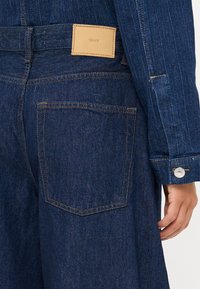 Asmuo, dėvintis tamsiai mėlynus džinsus ir atitinkančią džinsinę striukę, demonstruojantis galinį kišenę ir striukės rankovę su mygtukų užsegimu.