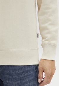 Sweatshirt couleur crème en tissu texturé, avec des poignets et un ourlet côtelés, et une petite étiquette de marque sur le côté.