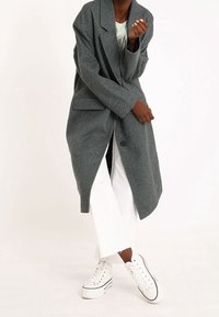 Lungo cappotto grigio in materiale texturizzato con colletto a revers, chiusura a un bottone e tasche laterali, abbinato a pantaloni bianchi e sneakers.