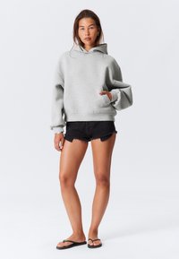 Gray hooded sweatshirt with a front pocket, paired with black denim shorts with frayed edges and black flip-flops, standing against a plain background.