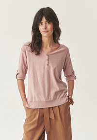 Maglietta a maniche lunghe rosa pallido con scollatura a V, tre bottoni in legno e polsini arrotolati. Tessuto morbido con orlo a costine. Indossata con pantaloni marroni.