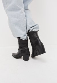 Bottes noires en cuir jusqu'à la cheville avec un bout légèrement pointu, un design texturé et un talon carré de hauteur moyenne. Portées avec un jean en denim bleu clair.