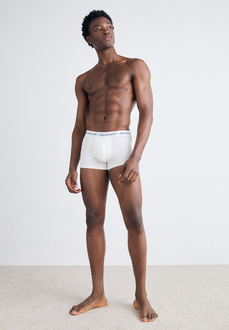 Boxer in cotone bianco con elastico in vita che presenta il nome del marchio "GANT" in blu. Il modello sta in piedi a piedi nudi su un tappeto chiaro.
