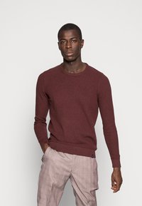 Bordeauxfarbener Strickpullover mit Rundhalsausschnitt, gerippten Bündchen und Saum. Getragen mit hellen Karohosen, die einen strukturierten Stoff zeigen.