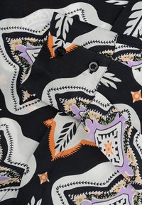 Tela negra con un patrón floral en blanco, naranja y púrpura. Presenta un cuello, dos botones negros y detalles de costura visibles.