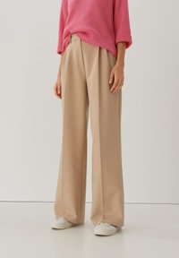 Pantalons larges beiges avec une texture lisse, taille haute et coupe décontractée, associés à des chaussures de sport blanches et un pull rose en côtes.